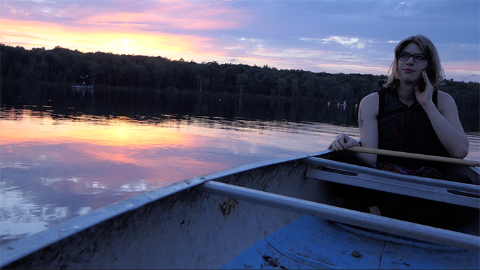 Zefyr, a white trans femme sits in a canoe on a lake at sunset. Her left hand is at her face as she looks off camera left. She has a slight smile. The sky behind her is pinks, yellows and blues and is reflected in the lake's surface.